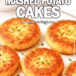 A close-up image of Crispy Mashed Potato Cakes garnished with chopped chives, arranged on a white marble board. A bowl of mashed potatoes, a bunch of fresh chives, and a neutral towel appear in the soft-focus background.