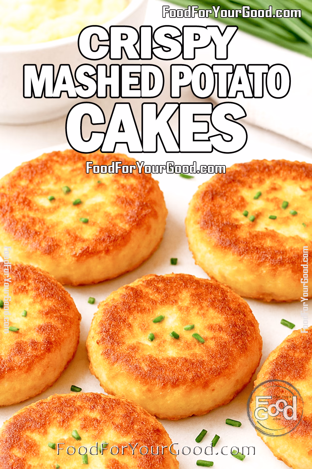 Crispy Mashed Potato Cakes on FoodForYourGood.com A close-up image of Crispy Mashed Potato Cakes garnished with chopped chives, arranged on a white marble board. A bowl of mashed potatoes, a bunch of fresh chives, and a neutral towel appear in the soft-focus background.