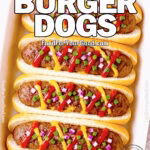 Cheesy Baked Burger Dogs topped with ketchup, mustard, red onion, and chives in a ceramic baking dish on a linen towel.
