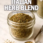 A small glass jar filled with homemade Classic Italian Herb Blend on a marble countertop, with a tarnished spoon of seasoning beside it.