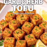 Golden crispy garlic herb tofu cubes served on a square white plate with lemon wedges and fresh parsley on a brown marble background.