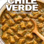 A white Dutch oven filled with fork-tender pork chile verde in a thick green sauce, garnished with cilantro, with a wooden spoon and white towel beneath.