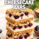 Blueberry Crumble Cheesecake Bars stacked on a marble surface, with a golden oat crust, creamy cheesecake layer, juicy blueberries, and streusel topping. Styled with fresh blueberries, oats, and a white towel in the background.