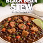 Brazilian Black Bean Stew in a white ceramic bowl with smoky sausage, tender pork, glossy black beans, colorful peppers, cilantro, and lime, styled on a white marble surface with rice and fresh peppers blurred in the background.