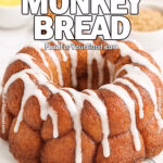 Freshly baked Caramel Cinnamon Monkey Bread drizzled with powdered sugar glaze on a white plate, set on a marble countertop with cinnamon sticks, melted butter, and brown sugar in the background.