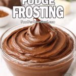 Chocolate Fudge Frosting in a glass bowl on white marble with cocoa powder, chocolate, and butter in the background.