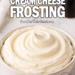 A creamy swirl of Classic Cream Cheese Frosting in a white bowl, set on a marble countertop with butter, vanilla, and a white kitchen towel in the background, photographed for FoodForYourGood.com