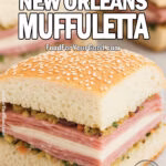 Classic New Orleans Muffuletta sandwich wedge with layers of meats, cheeses, and olive salad on a sesame loaf, served on marble with more sandwiches blurred in the background.