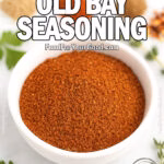 White ceramic bowl filled with Classic Old Bay Seasoning, surrounded by fresh parsley, thyme sprigs, and small piles of colorful spices on a marble countertop.
