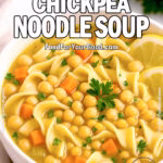 A bowl of Cozy Chickpea Noodle Soup with egg noodles, carrots, chickpeas, and parsley, served with lemon slices on a white countertop.