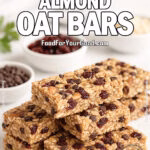 Close-up portrait of stacked Cranberry Almond Oat Bars on a white marble countertop, with bowls of cranberries, almonds, and chocolate chips blurred in the background, at FoodForYourGood.com