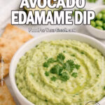 Homemade-style avocado edamame dip in a white bowl with rustic texture, topped with cilantro, on marble with pita chips.