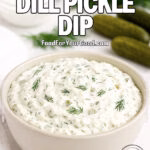 A creamy dill pickle dip in a beige bowl topped with fresh dill, with pickles, dill sprigs, and sour cream blurred in the background on a white marble surface.