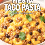 Creamy Fiesta Taco Pasta served in a beige ceramic bowl with elbow macaroni, ground beef, black beans, and corn in a cheesy sauce, garnished with cilantro on a white marble background with fresh ingredients blurred behind.