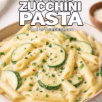 A bowl of creamy penne pasta with zucchini slices, garnished with fresh parsley and Parmesan, on a white marble surface with fresh ingredients in the background.