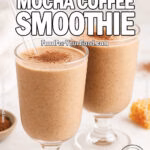 Two footed glasses filled with creamy mocha coffee smoothie, each with a wide glass straw, set on a white marble surface with cocoa powder, honeycomb, and a white towel in the blurred background.