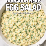 Overhead view of Creamy Tarragon Herb Egg Salad in a white bowl with fresh herbs and lemon on marble at FoodForYourGood.com