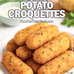 Crispy Garlic Potato Croquettes stacked on a white plate with chives, Parmesan, and potatoes on a marble background.