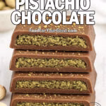 Rustic Dubai Pistachio Chocolate bars stacked on white marble, filled with textured pistachio cream and dusted with chopped pistachios, with whole pistachios scattered in front and a blurred bowl in the background.