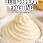 A bowl of freshly made Ermine Buttercream Frosting with a soft, slightly textured swirl, placed on a white marble countertop, with a piping bag, vanilla cupcakes, milk, and sugar blurred in the background.