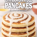 A stack of Fluffy Cinnamon Roll Pancakes drizzled with cream cheese glaze, served on a white plate over marble with blurred cinnamon sticks, butter, and brown sugar in the background.
