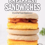 Two stacked Freezer Breakfast Sandwiches on toasted English muffins, filled with baked eggs, melted cheddar cheese, and bacon or sausage. Placed on white marble with a plain white wall background and herbs blurred on the side.