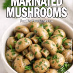 White ceramic bowl filled with marinated mushrooms garnished with fresh parsley, set on a white marble countertop with herbs and a white towel in the blurred background.