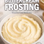 Homemade German Buttercream Frosting in a white bowl with spoon, cupcakes, and piping bag on marble background.
