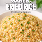A white bowl filled with golden garlic fried rice garnished with green onions, set on a white marble surface with fresh herbs blurred in the background.