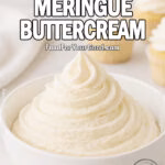 Italian Meringue Buttercream in a white ceramic bowl on a marble surface with frosted cupcakes in the blurred background.