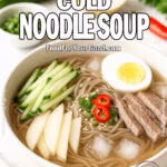 Bowl of Korean Cold Noodle Soup with soba noodles, cucumber, pear, beef slices, boiled egg, and chili in icy broth on a marble surface.