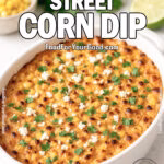 Mexican Street Corn Dip baked in a white dish, topped with Cotija cheese and cilantro, on marble with lime and corn.