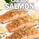 Parmesan Caesar Salmon served on a white plate with a crispy golden crust, garnished with parsley and lemon wedges, on a white marble background with Caesar dressing in the background.
