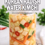 Refreshing Korean Radish Water Kimchi in a glass jar with crisp radish, napa cabbage, carrots, green onion, and chili in a clear brine, styled on a white marble background with fresh herbs and towel.