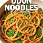 A bowl of sesame garlic udon noodles with carrots, snow peas, and fresh herbs on a white marble surface.
