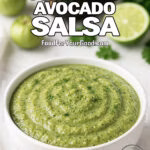 A bowl of Smoky Tomatillo Avocado Salsa on a white marble countertop, with fresh tomatillos, avocado, lime, cilantro, and a white towel in the background.