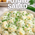 A rustic ceramic bowl filled with sour cream and chive potato salad, garnished with fresh chives, surrounded by whole baby potatoes and herbs on a marble surface.