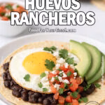 Sunrise Huevos Rancheros with crispy tortilla, black beans, sunny-side-up egg, fresh salsa, avocado slices, and queso fresco on a white plate over marble.