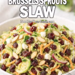 Sweet & Tangy Brussels Sprouts Slaw in a white ceramic bowl, topped with almonds, cranberries, and feta. Styled on a marble surface with fresh Brussels sprouts and a white towel in the background.