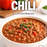 Texas-Style Chili in a white ceramic bowl on a white marble countertop, garnished with fresh cilantro and surrounded by bell peppers, onion, and herbs in the background.