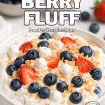 Whipped Berry Fluff in a white bowl topped with fresh strawberries, blueberries, marshmallows, graham cracker crumbs, and toasted coconut on a marble background.