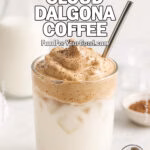 Whipped Cloud Dalgona Coffee in a clear glass with a messy whipped topping, straw inserted, served on a white marble countertop with blurred milk bottle, cocoa powder, ice cubes, and a white towel in the background on FoodForYourGood.com
