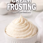 Whipped Cream Frosting in a white ceramic bowl on a marble countertop, surrounded by blurred ingredients like cream, sugar, and vanilla.