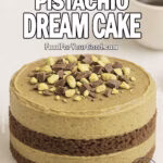 Chocolate Pistachio Dream Cake layered with cocoa sponge and pistachio chocolate frosting, topped with chopped pistachios and shaved chocolate on a white marble background.