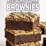 A stack of fudgy Dubai Pistachio Tahini Brownies on a round white plate. The brownies feature a rich chocolate base, a smooth pistachio-tahini layer, and are topped with sesame seeds, chopped pistachios, and a drizzle of dark chocolate. Styled on a white marble surface with a blurred white towel in the background.