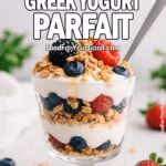 A homemade honey berry Greek yogurt parfait served in a short-stem glass with layers of creamy yogurt, granola, and fresh strawberries, blueberries, and blackberries. Topped with honey drizzle and a modern silver spoon on a white marble background, surrounded by berries and a blurred white towel for an elegant, natural look.