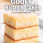 St. Louis Gooey Butter Cake squares stacked on a white marble tray with a soft, fluffy cake base and a slightly uneven baked cream cheese top, dusted with powdered sugar. A white baking pan with more cake is blurred in the background, styled with a white towel and powdered sugar bowl